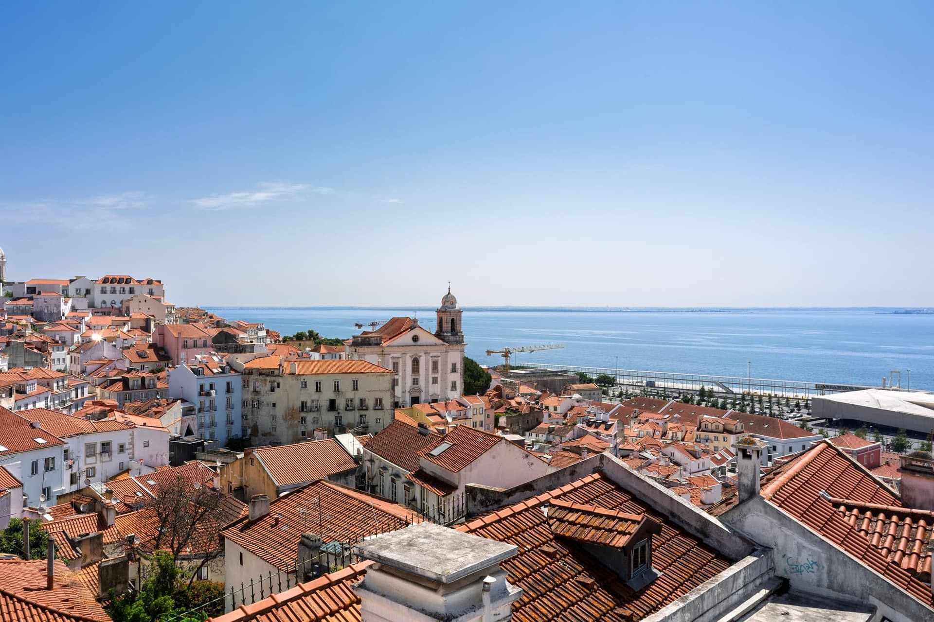 Terracotta rooftops of Lisbon's Alfama district stepping down towards the wide grey-blue Tagus river.
