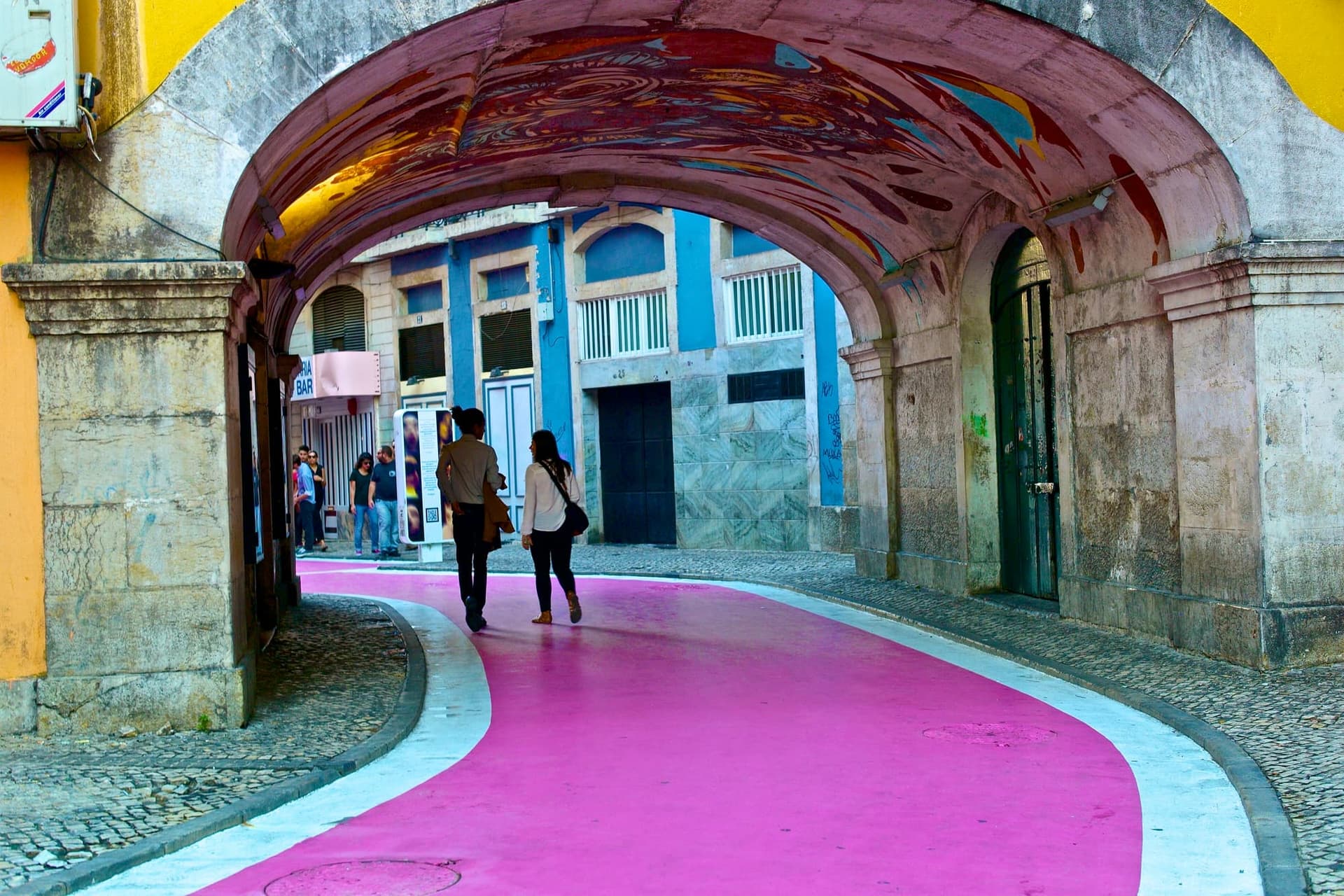 A narrow Lisbon street painted bright pink underfoot, with bars on both sides and umbrellas strung overhead.