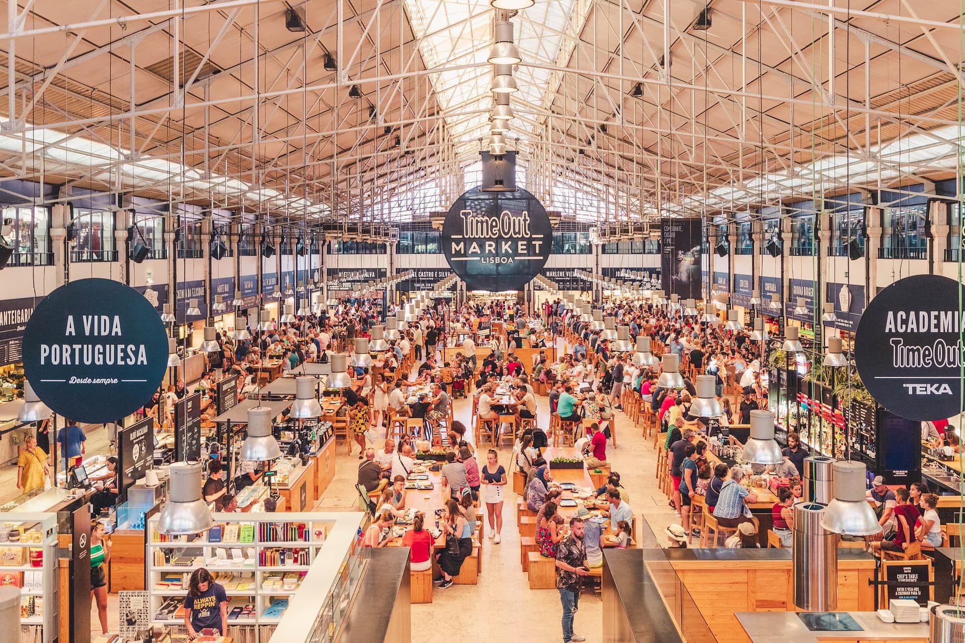 Long communal tables filling the hall of a covered food market in Lisbon, with food counters along the walls.