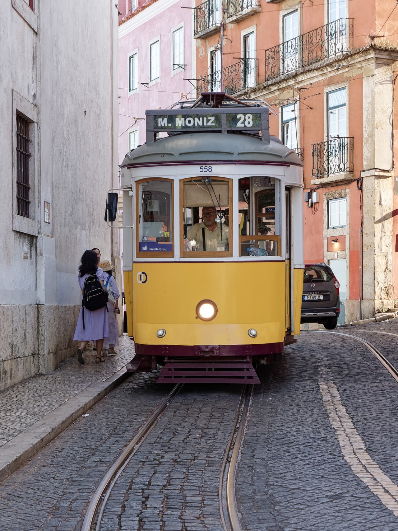 A yellow number 28 tram waiting on a narrow cobbled street beside a tiled wall in Lisbon.