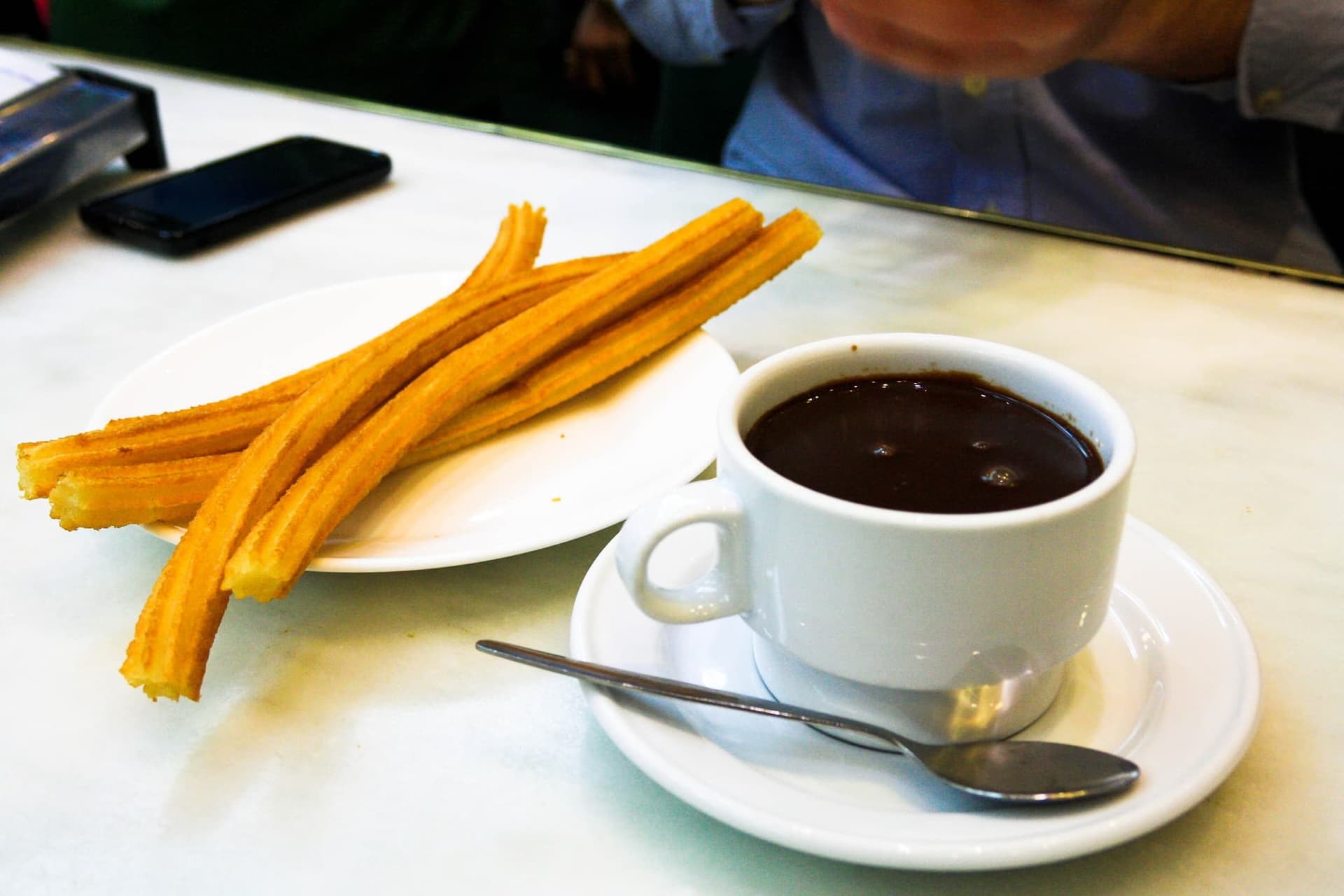 A plate of churros beside a thick cup of drinking chocolate on a marble table.