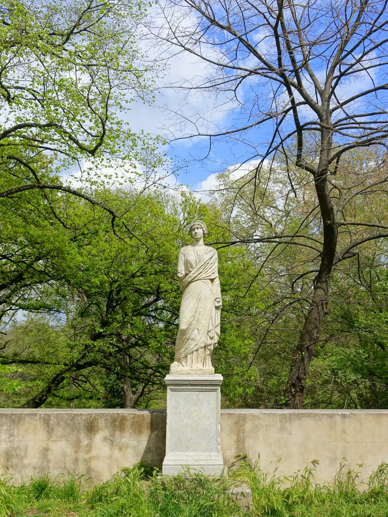 A weathered stone statue standing among the pines and columns of the Villa Borghese gardens in Rome.
